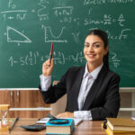 young female teacher sits at table with school tools raising pen in hand
