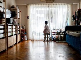 Child is standing alone in front of the window of the house