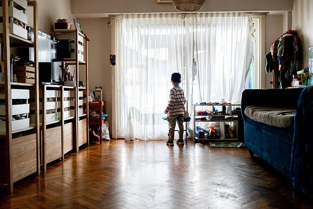 Child is standing alone in front of the window of the house